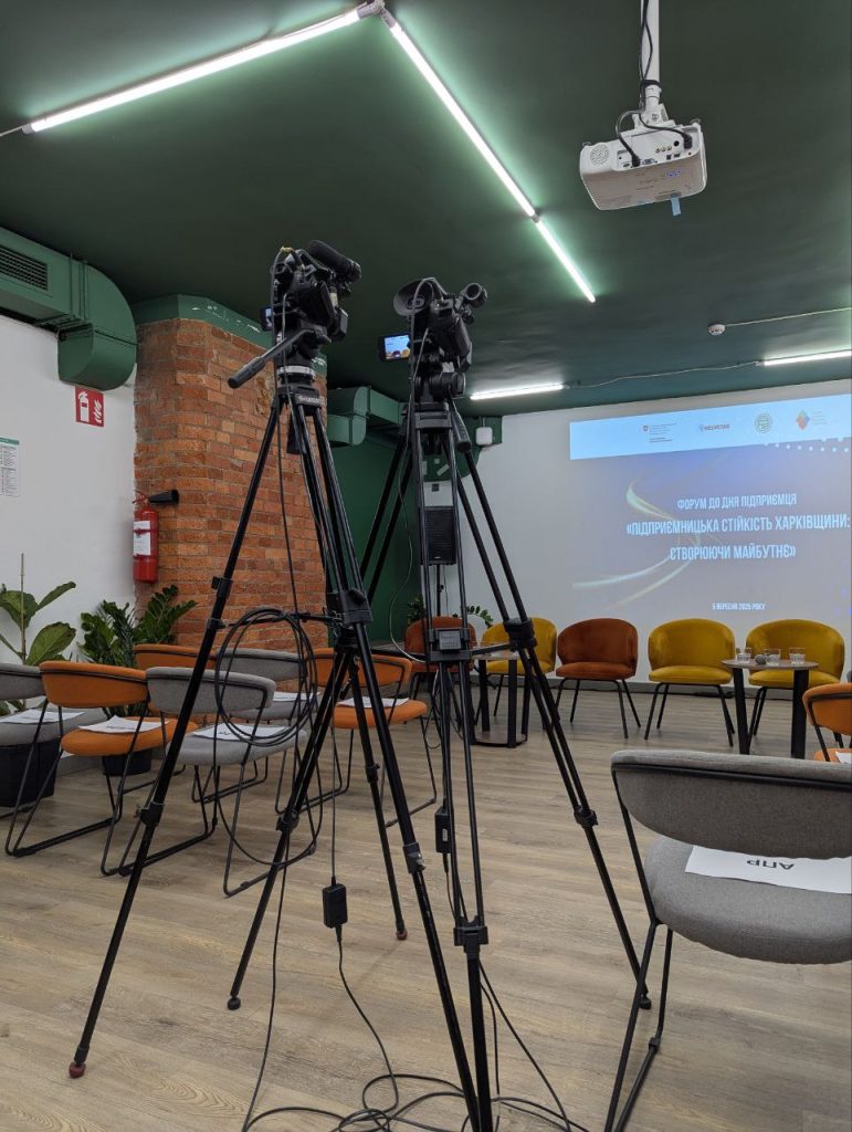 11 Порожня зала пресцентру «Накипіло» перед початком заходу. У проході між кріслами слухачів стоять камери на штативах, позаду крісел спікерів знаходиться екран, на якому відображається напис «Форум до Дня підприємця "Підприємницька стійкість Харківщини: створюючи майбутнє"».