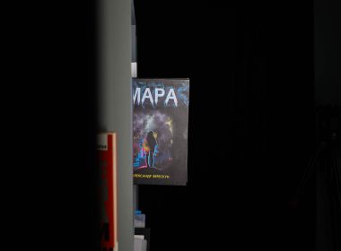 Книга «Мара» Олександра Верескуна виглядає з-за краю книжкової шафи в темній кімнаті; видно частину обкладинки з назвою, силуетом людини й ім'ям автора.