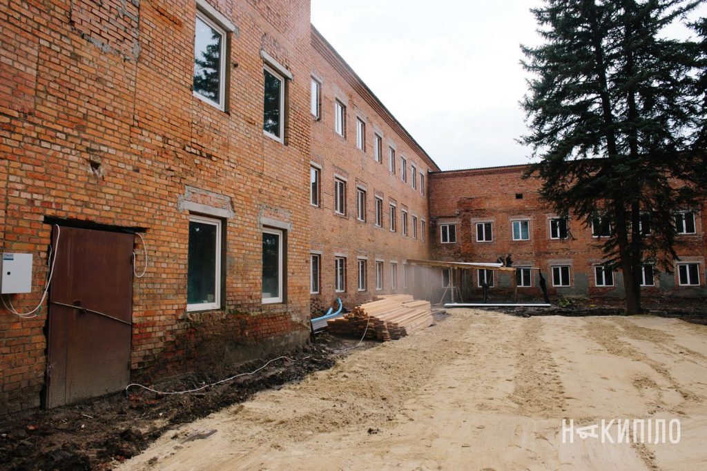 У Змієві реконструюють будинок під житло для ВПО: деталі проєкту Внутрішнє подвір’я цегляної будівлі, де тривають будівельні роботи. На землі лежать дошки та будівельні матеріали, частину території засипано піском. Житло.