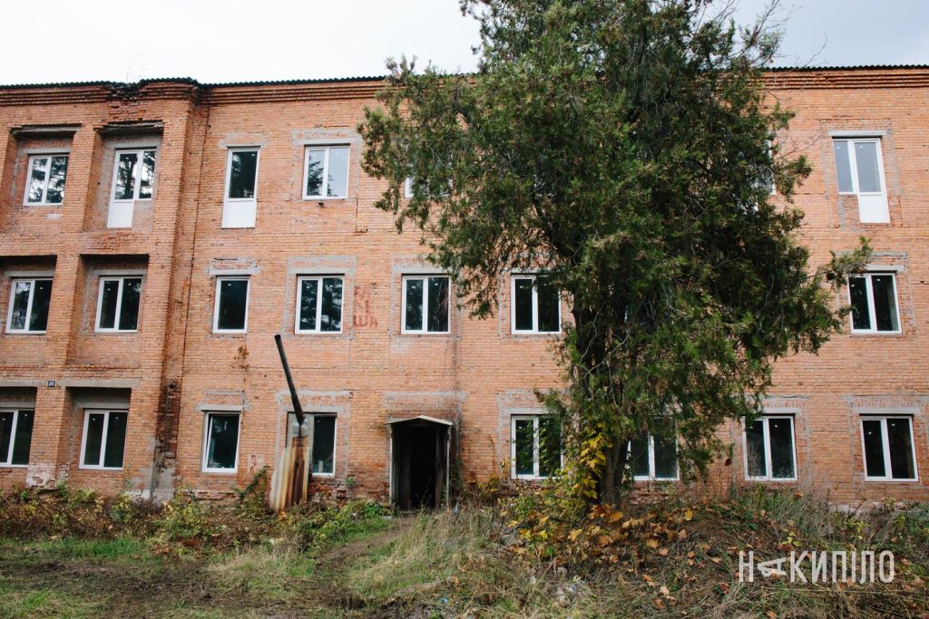 У Змієві реконструюють будинок під житло для ВПО: деталі проєкту Фасад триповерхової цегляної будівлі з установленими пластиковими вікнами. Перед входом росте велике хвойне дерево. Житло.